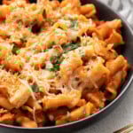 A black bowl with chicken parmesan pasta with fresh herbs and cheese on top with gold spoons on the side