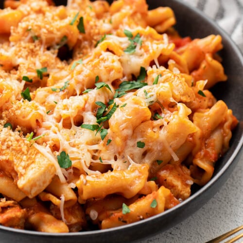 A black bowl with chicken parmesan pasta with fresh herbs and cheese on top with gold spoons on the side