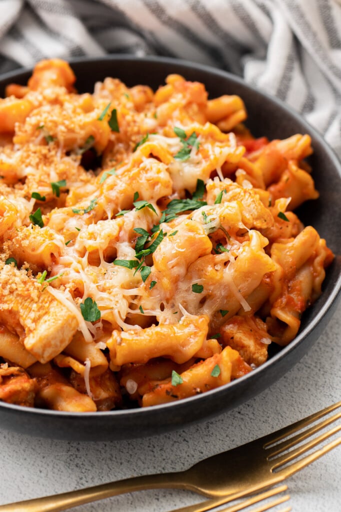 A black bowl with chicken parmesan pasta with fresh herbs and cheese on top with gold spoons on the side