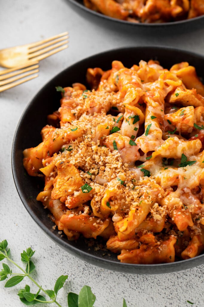 A black bowl with chicken parmesan pasta with fresh herbs and cheese on top with gold forks in the background