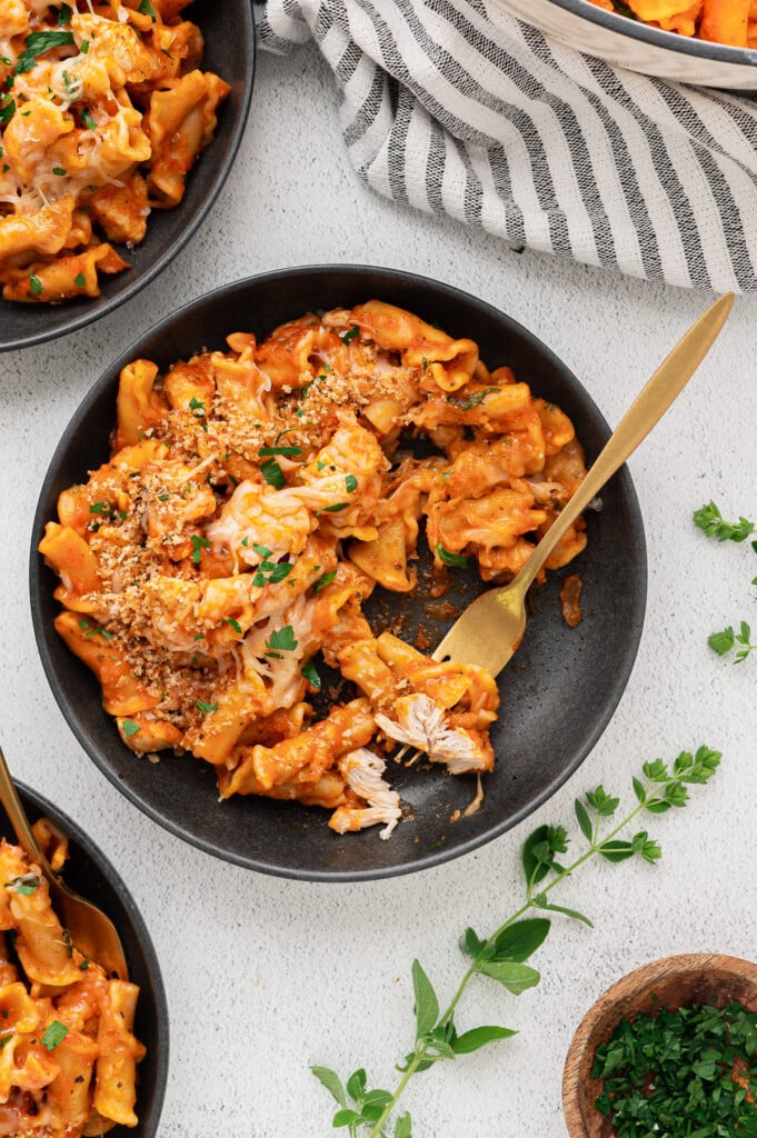 An overhead shot of a black bowl of chicken parm pasta with a gold fork taking a bite out of it with more bowls and herbs on the side