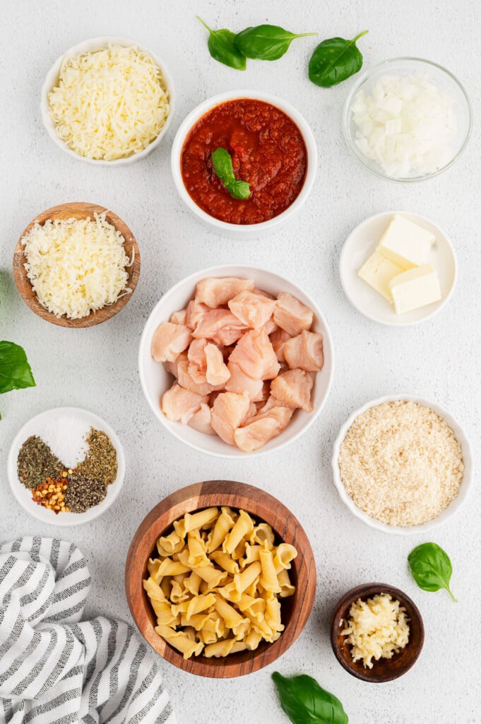 Ingredients for one pot chicken parm pasta in white bowls: butter, garlic, panko, pepper, olive oil, chicken breasts, onion, garlic, dried basil, dried oregano, red pepper flakes, campanelle pasta, marinara sauce, water, mozzarella cheese, parmesan cheese, fresh basil