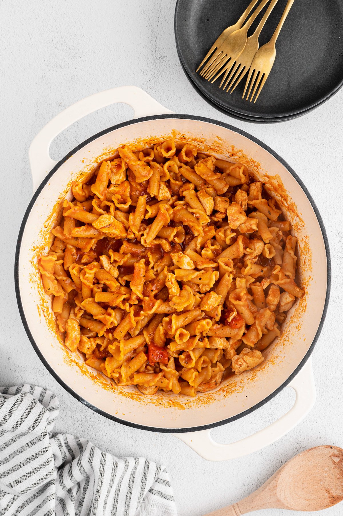 Chicken parmesan pasta in a dutch oven before the cheese is added on a white background with a towel, black bowls, and gold forks on the side