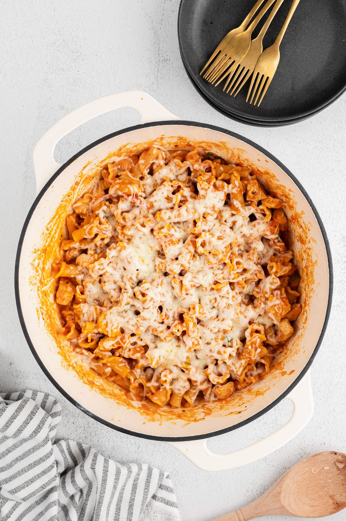 Chicken parmesan pasta in a dutch oven with cheese on top on a white background with a towel, black bowls, and gold forks on the side