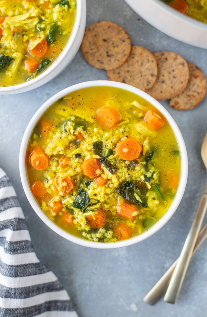 Chicken pastina soup in a white bowl on a grey counter with crackers and another bowl of soup on the side