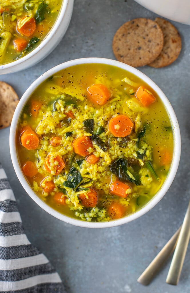 Chicken pastina soup in a white bowl on a grey counter with crackers and another bowl of soup on the side