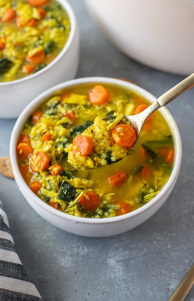 Chicken pastina soup in a white bowl with a spoon taking a scoop out of it on a grey counter with another bowl of soup in the background