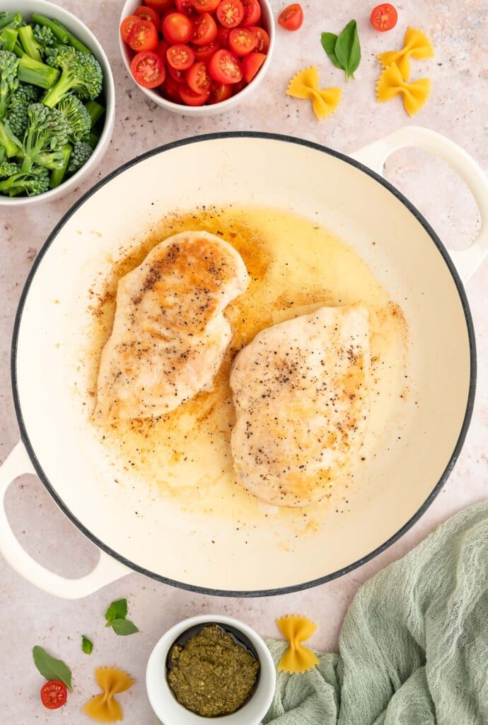 Two chicken breasts in olive oil in a dutch oven with more ingredients for chicken pesto pasta on the side.