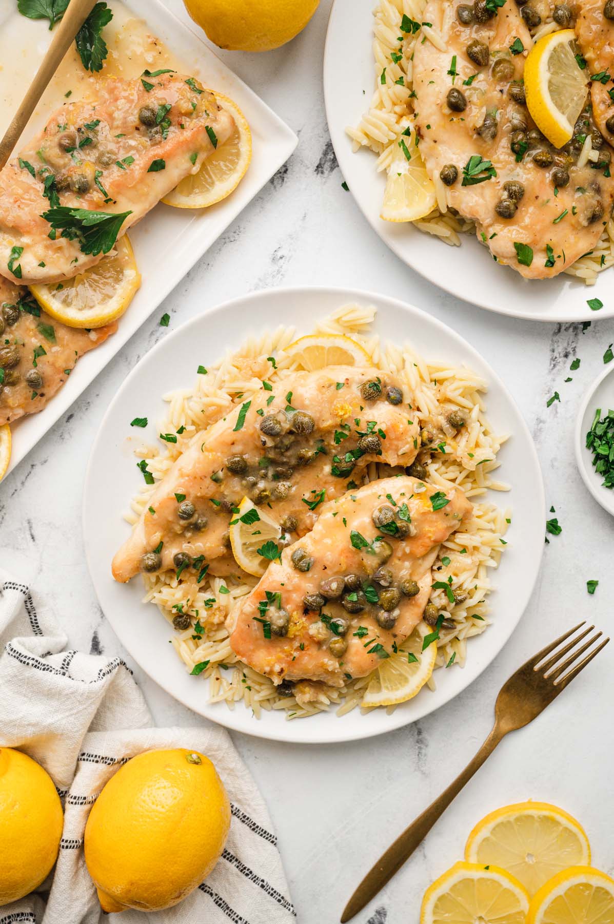 An overhead shot of chicken piccata on a white plate with a fork, lemons, another plate and platter on the side