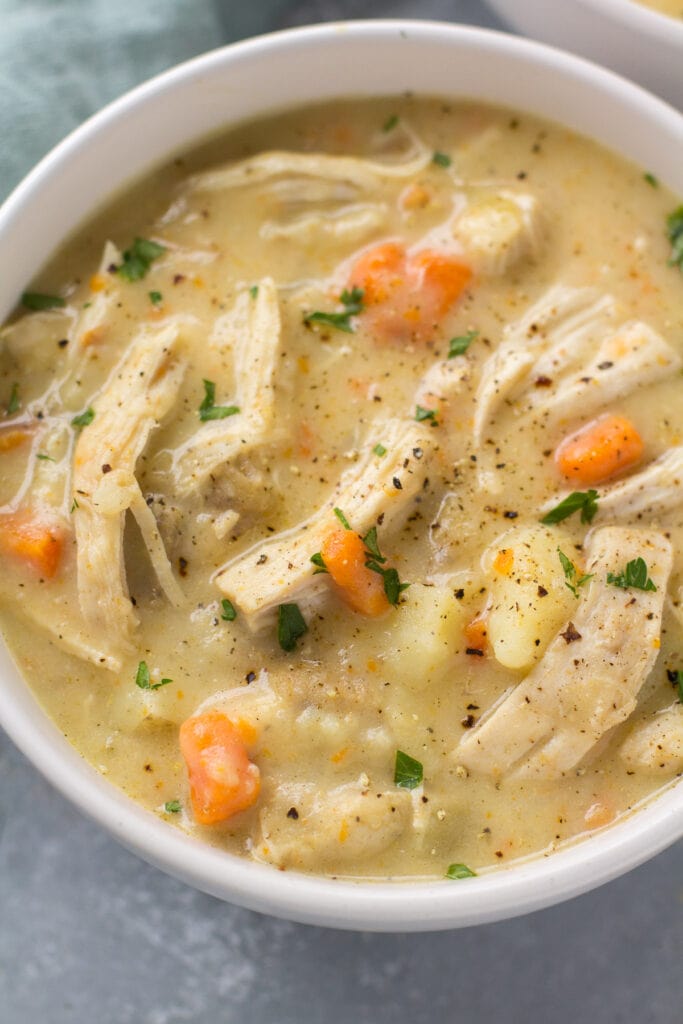 A close up shot of healthy chicken pot pie soup with black pepper on top in a white bowl. pieces of chicken, carrots and potatoes are peeking out of the bowl