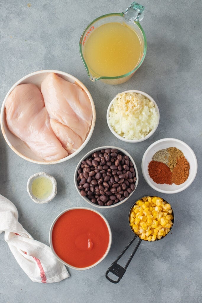 Ingredients for chicken taco soup in white bowls: olive oil, onion, garlic, chicken breasts, tomato sauce, chicken broth, paprika, pepper, chili powder, cumin, black beans, frozen corn