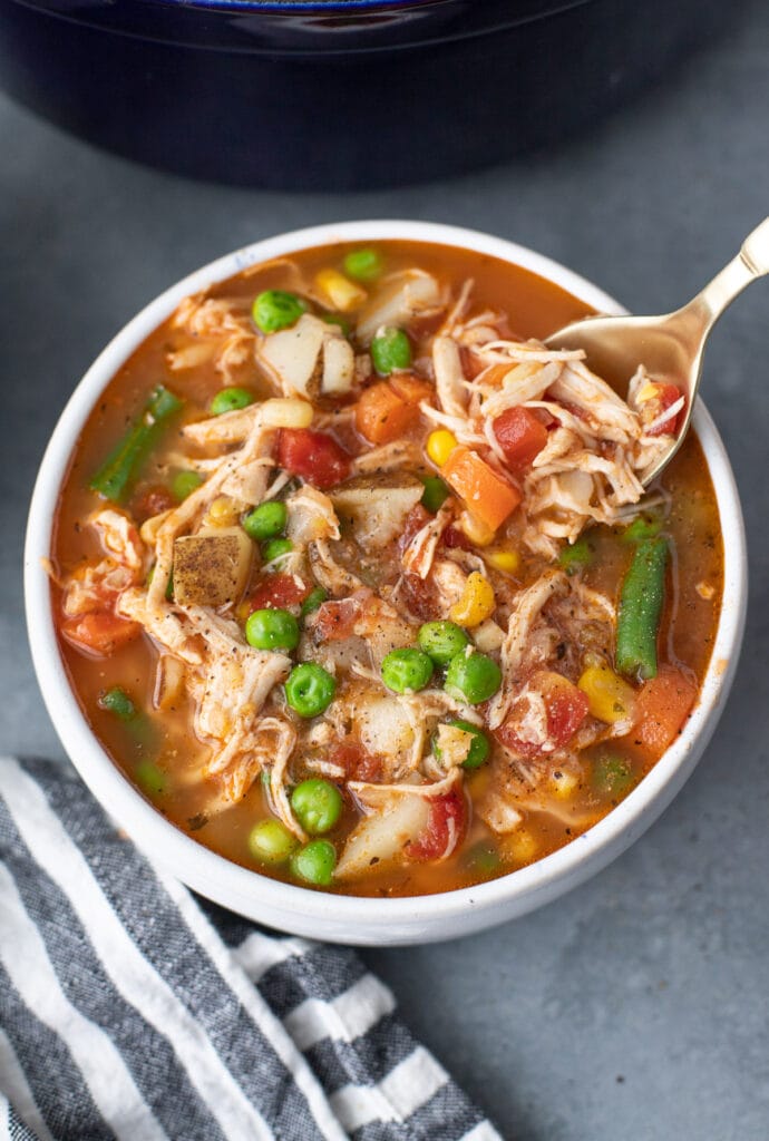 a white bowl chicken vegetable soup with a spoon taking a bite out of it on a grey background