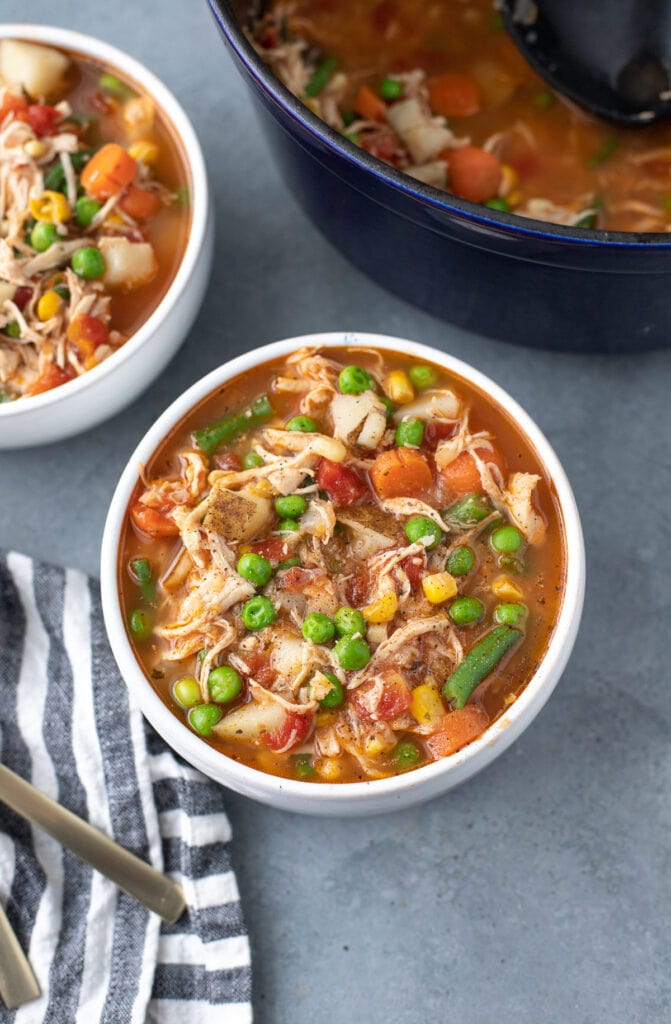 a bowl of chicken vegetable soup with another bowl and stock pot on the side on a grey background