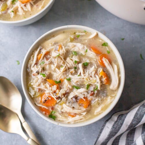An overhead shot of a white bowl of chicken and wild rice soup with another bowl of soup and spoons on the side