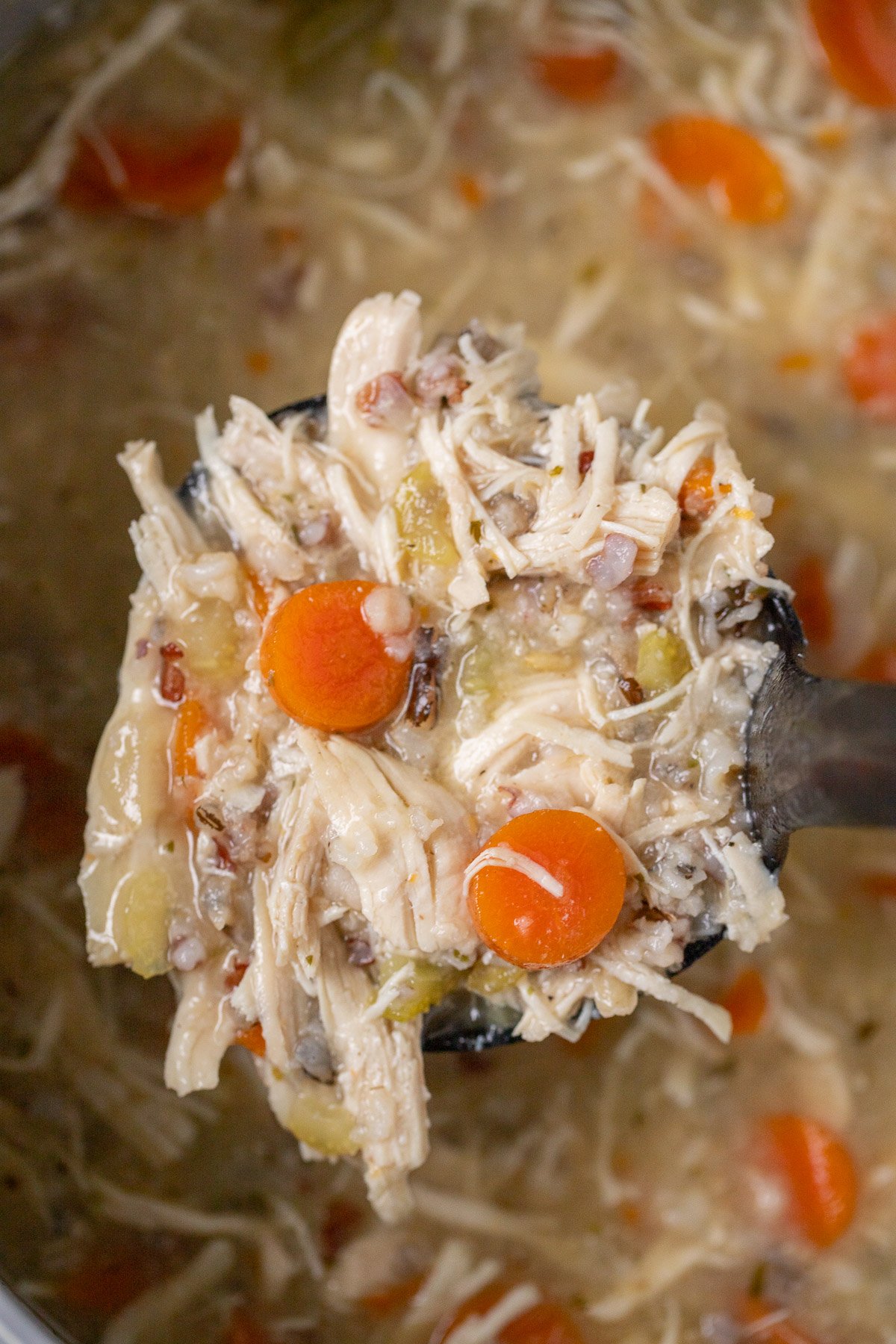 A close up of a ladle of chicken and wild rice soup with a pot of soup in the background