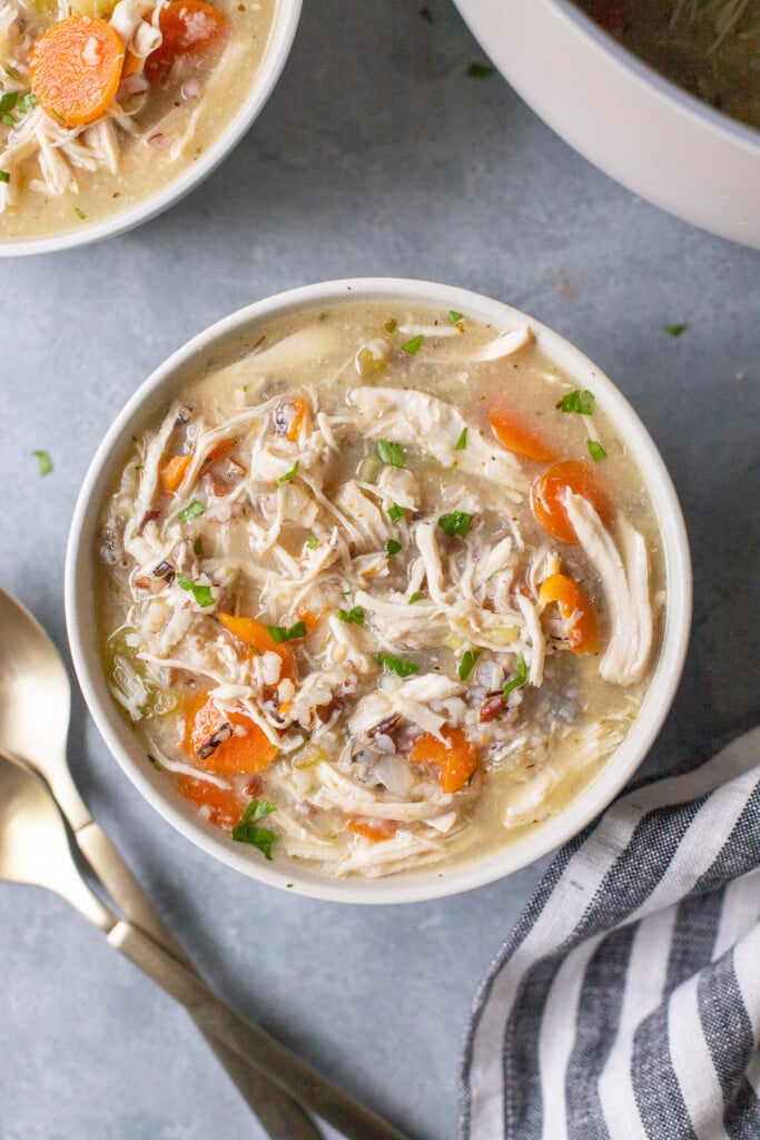 An overhead shot of a white bowl of chicken wild rice soup with another bowl of soup and spoons on the side
