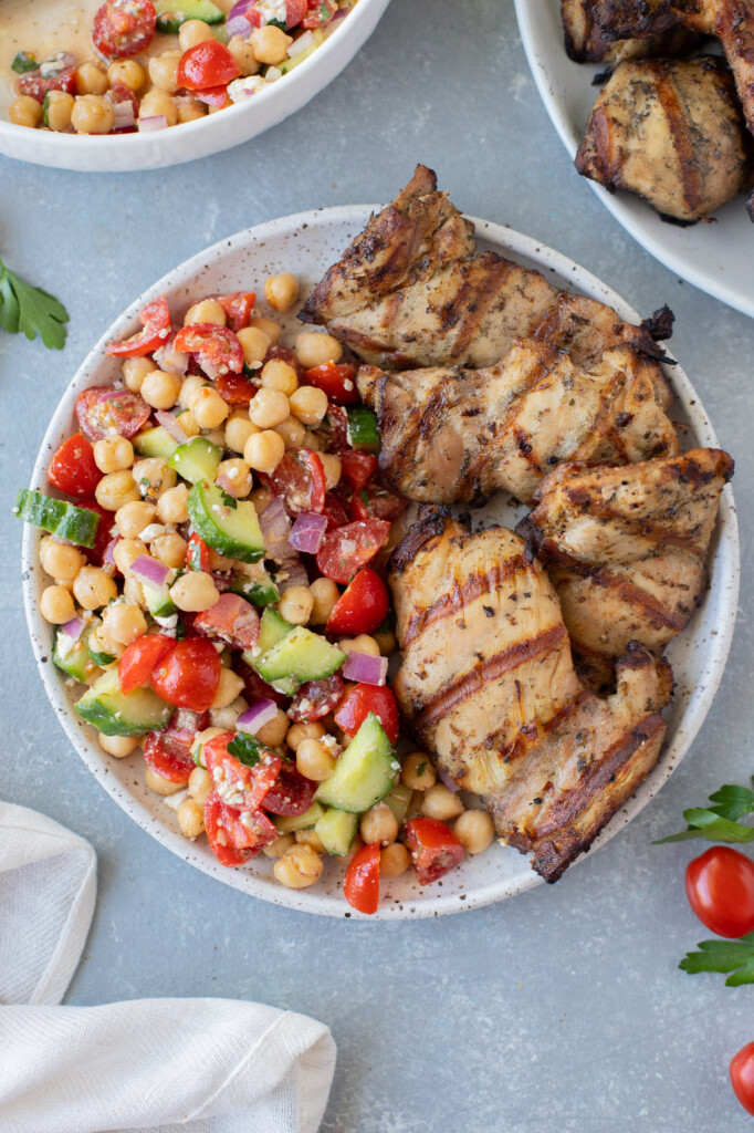 An overhead shot of a plate with greek chickpea salad on half with grilled chicken thighs on the other half on a grey background 
