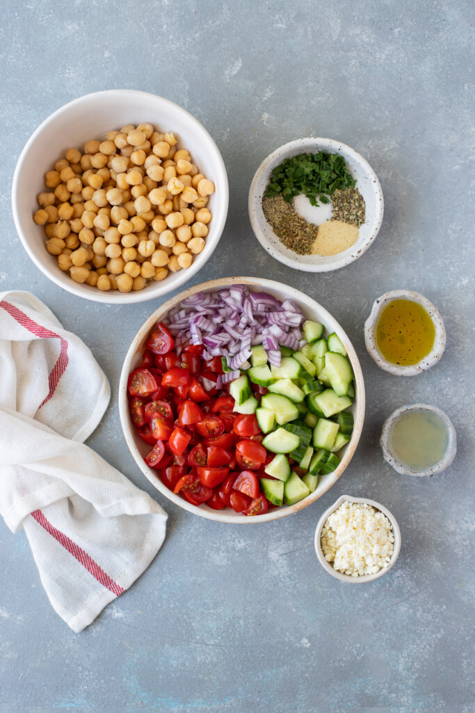 Ingredients for Mediterranean chickpea salad in small bowls: red onion, cherry tomatoes, cucumbers, chickpeas, olive oil, lemon juice, garlic powder, dried basil, dried oregano, salt, pepper, fresh parsley, crumbled feta