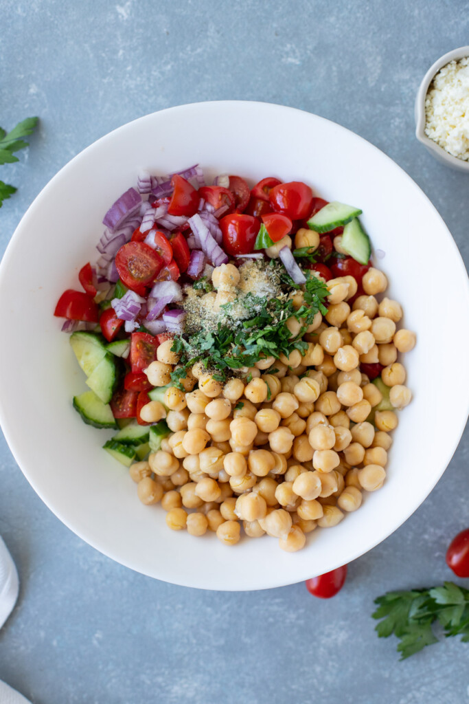 Ingredients for Mediterranean chickpea salad in a large white bowl before they have been mixed together on a grey background