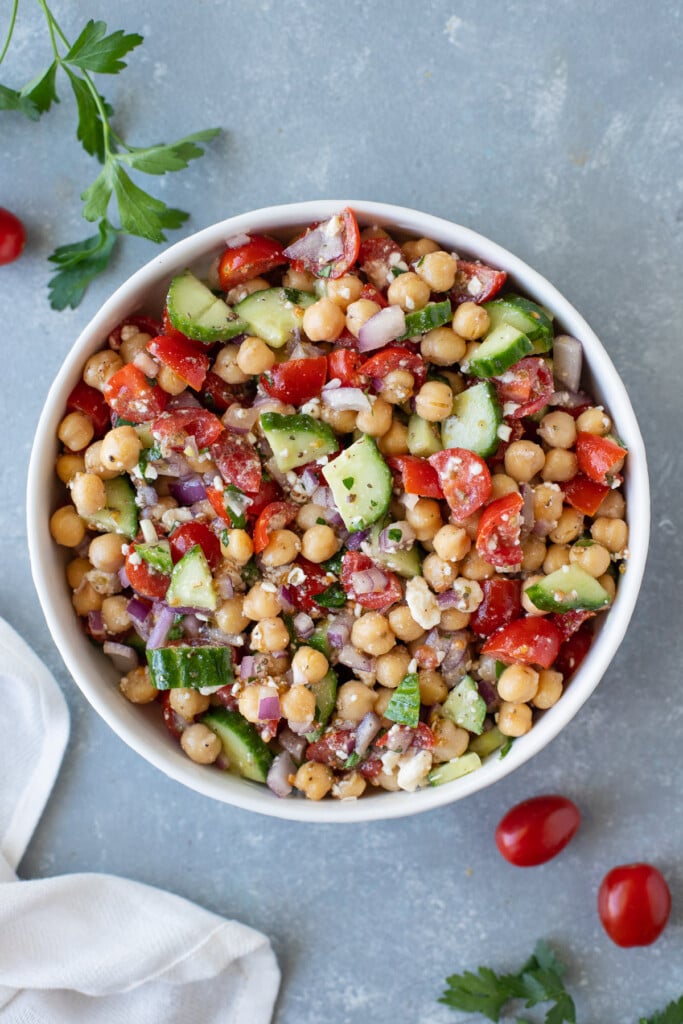 An overhead shot of a white bowl of Mediterranean chickpea salad on a grey background with tomatoes and herbs on the side