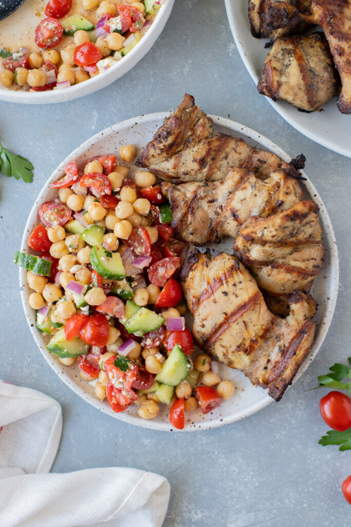 An overhead shot of a plate with greek chickpea salad on half with grilled chicken thighs on the other half on a grey background with a bowl of the salad and plate with chicken on the side