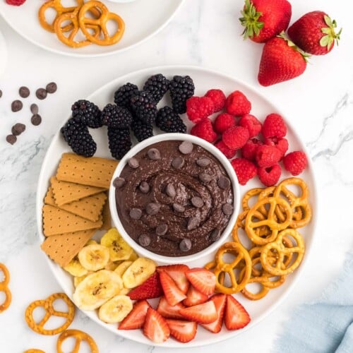 An overhead shot of a small bowl of chocolate hummus topped with chocolate chips surrounded by berries, graham crackers, and pretzels.