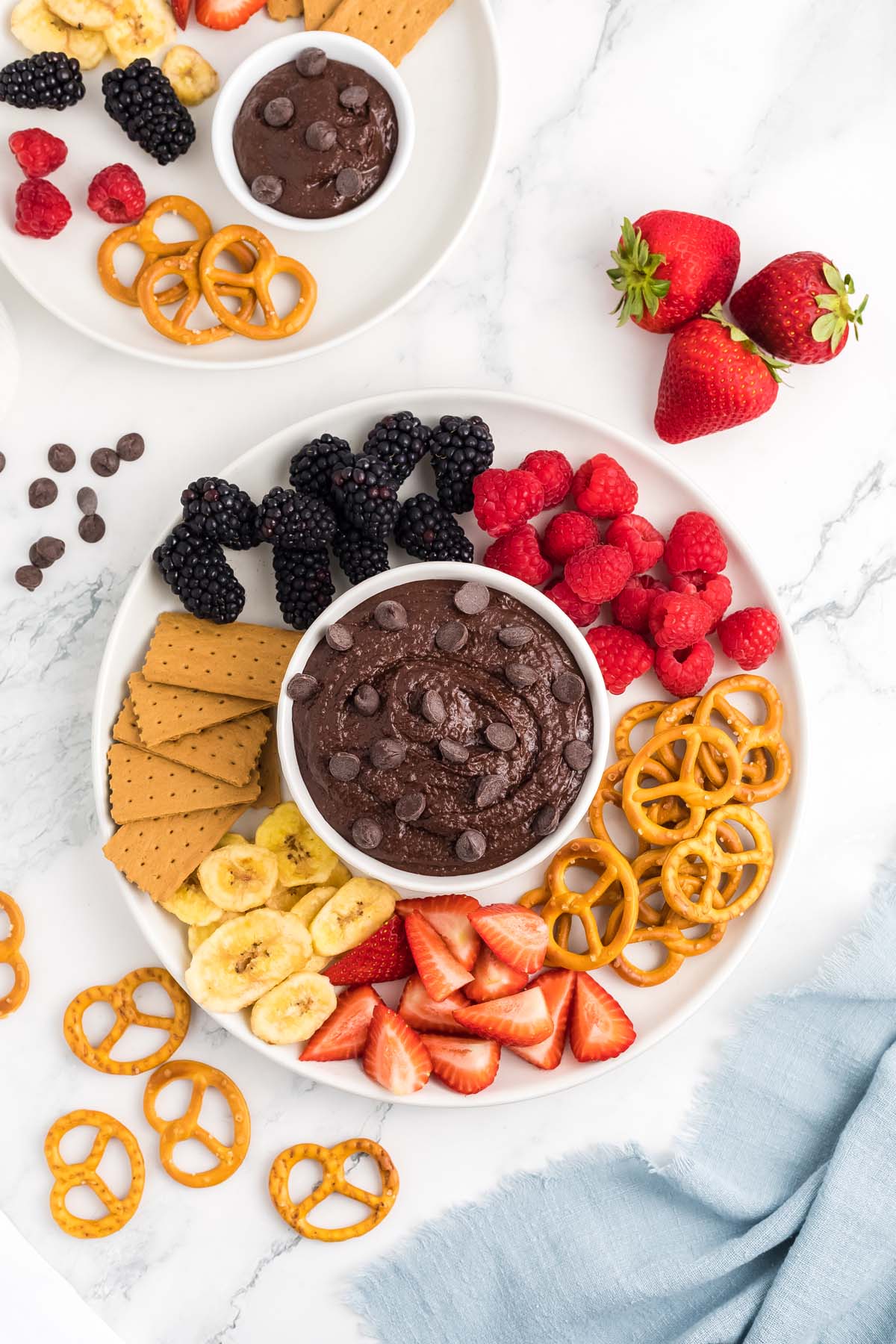 An overhead shot of a small bowl of chocolate hummus topped with chocolate chips surrounded by berries, graham crackers, and pretzels.
