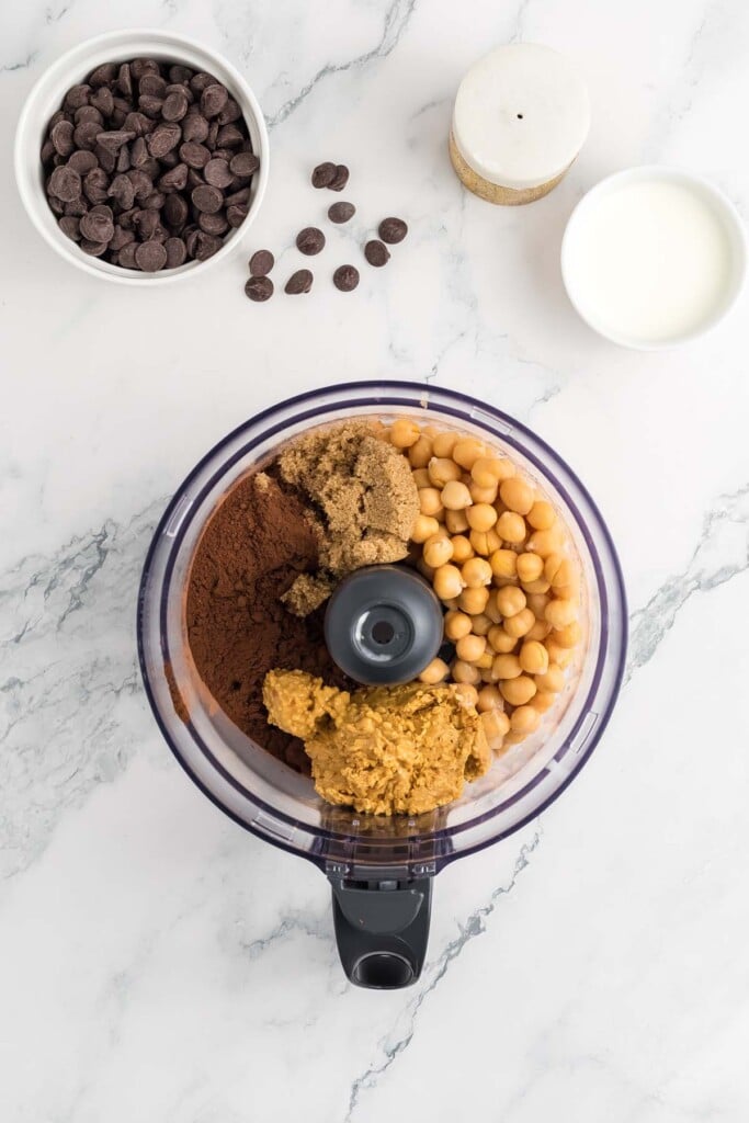 Ingredients for chocolate hummus in a food processor before blending with a small bowl of chocolate chips on the side.