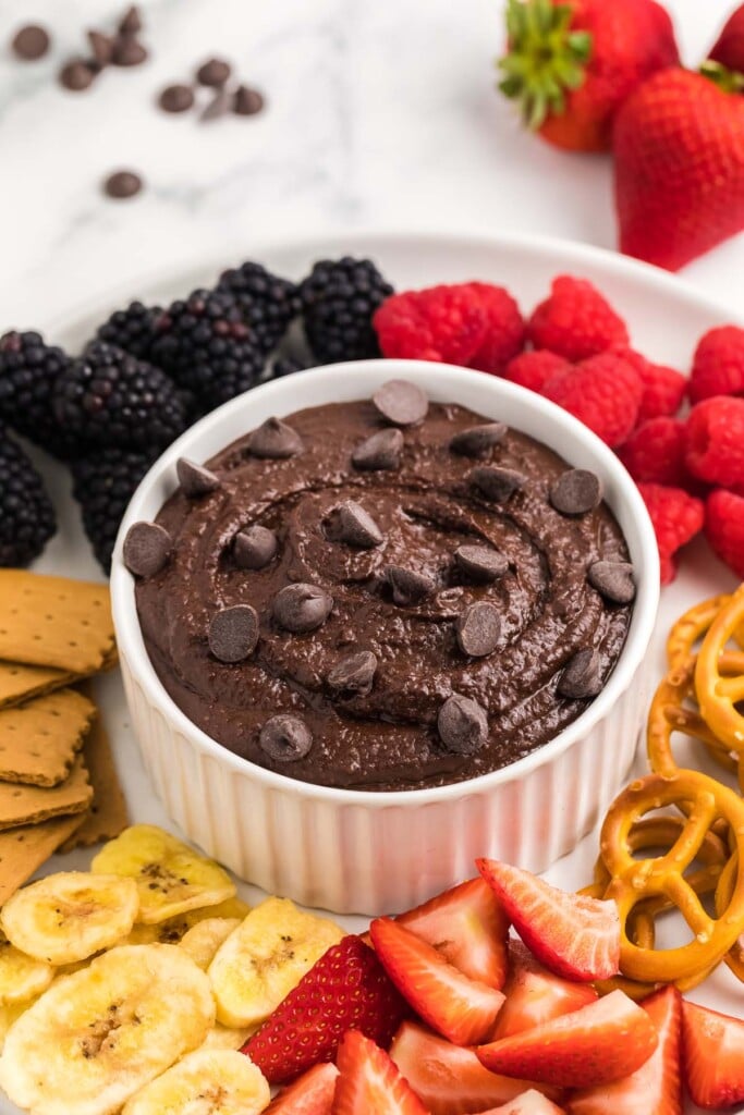 A small white bowl of chocolate hummus topped with chocolate chips with berries, banana slices, pretzels and graham crackers on the side.