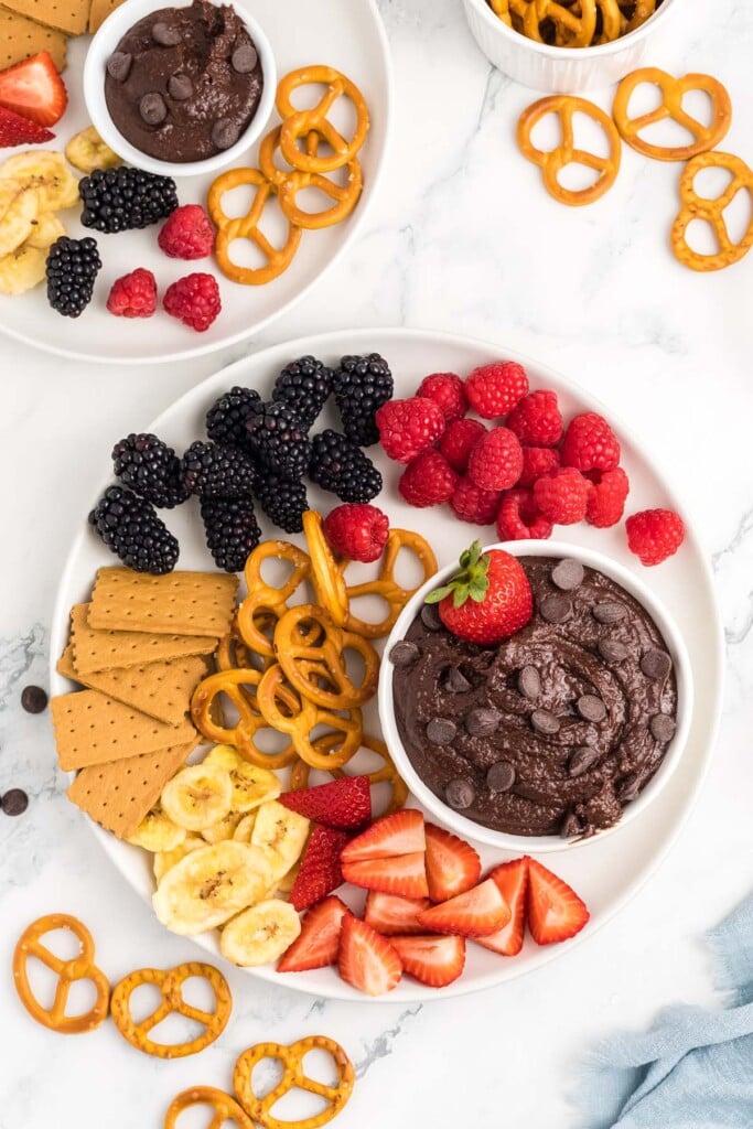 Chocolate hummus in a small white bowl topped with chocolate chips and a strawberry surrounded by blackberries, raspberries, strawberry slices, banana slices, pretzels, and graham crackers.
