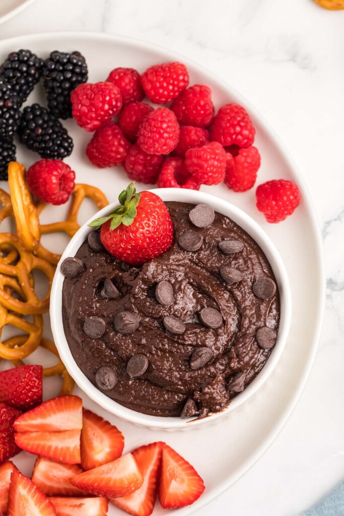 Chocolate hummus in a small white bowl topped with chocolate chips and a strawberry surrounded by blackberries, raspberries, strawberries, banana slices, and pretzels.