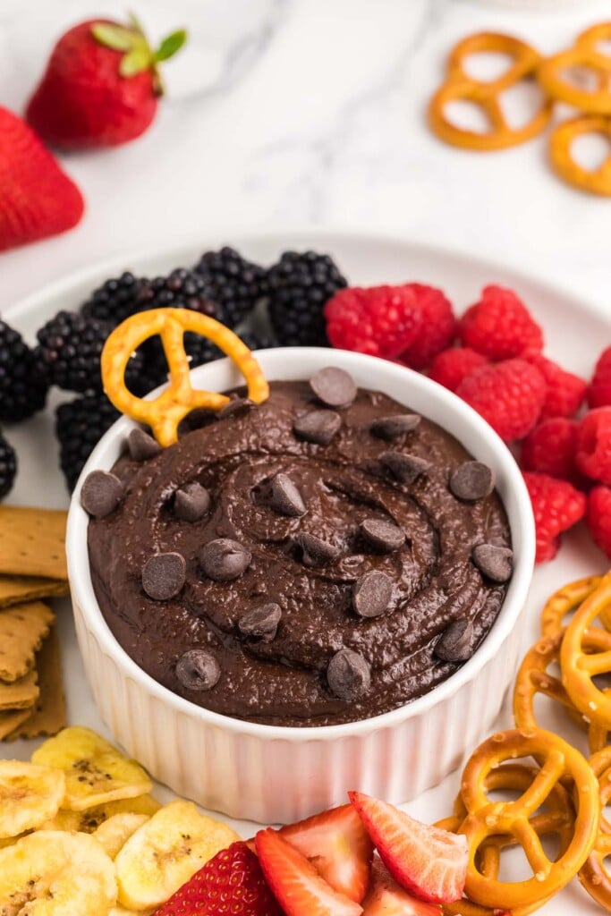 Chocolate hummus in a small white bowl topped with chocolate chips and a pretzel dipped in it surrounded by blackberries, raspberries, strawberries, banana slices, pretzels, and graham crackers.