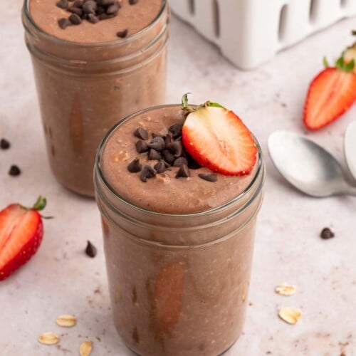 Two mason jars with chocolate overnight oats topped with mini chocolate chips and sliced strawberries.
