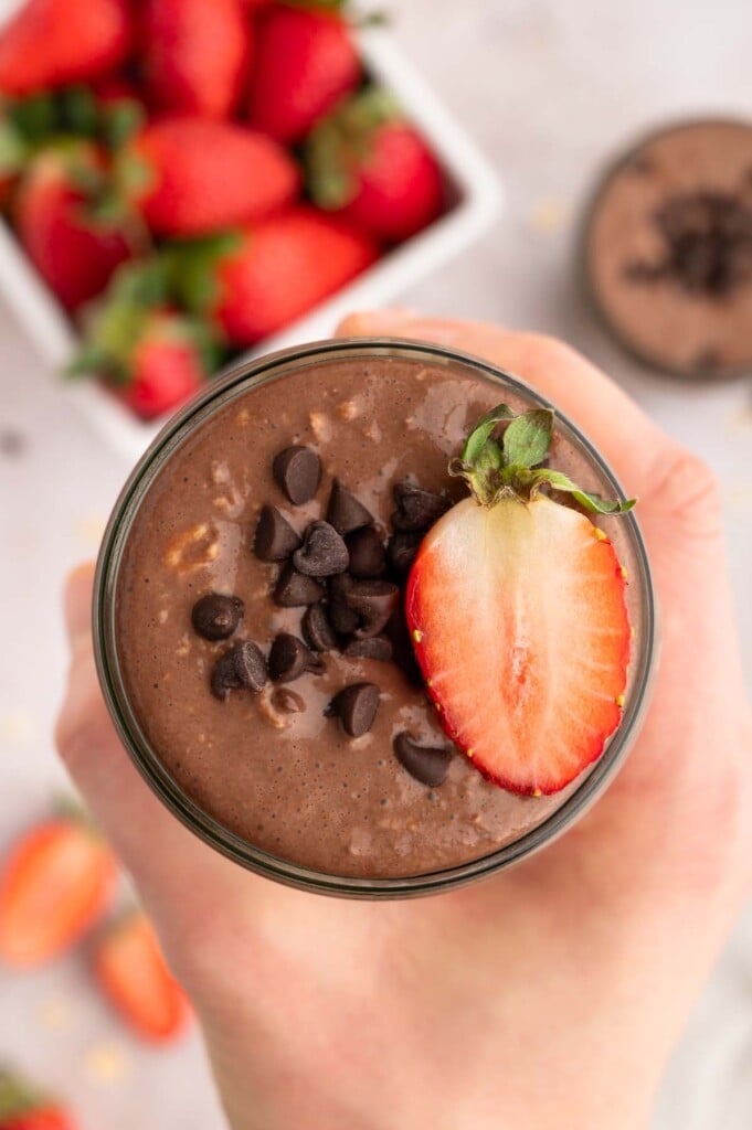 A hand holding up a mason jar of chocolate overnight oats topped with mini chocolate chips and a sliced strawberry.