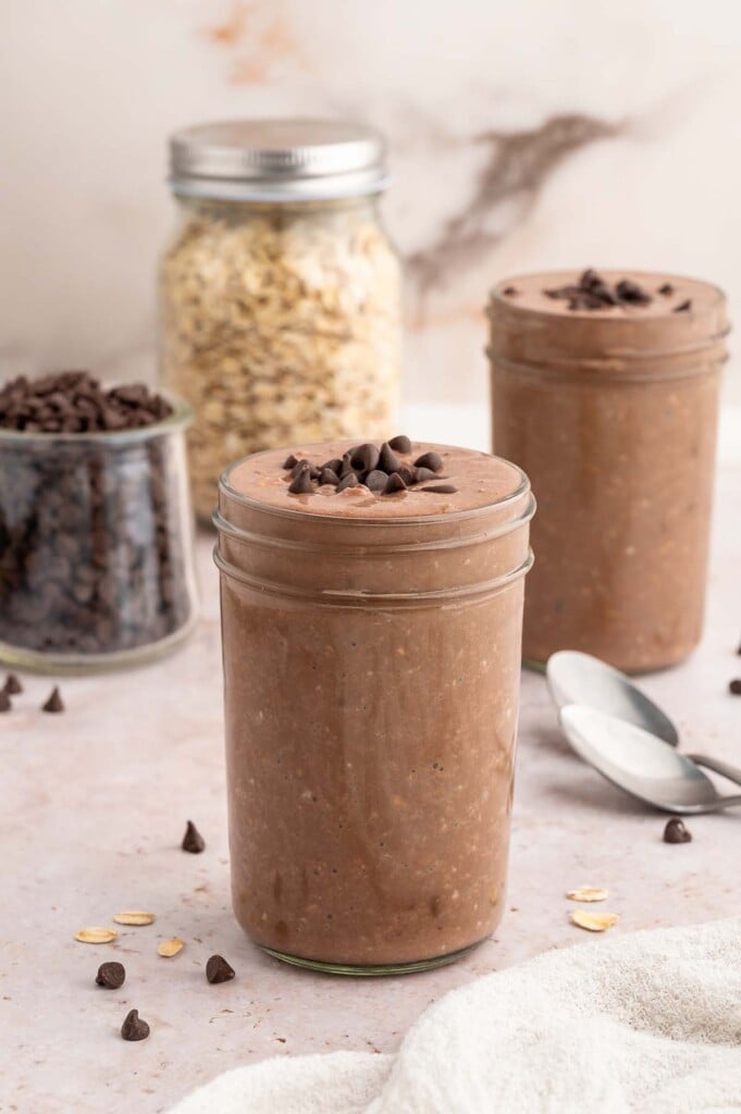 Two mason jars with chocolate overnight oats topped with mini chocolate chips and more chocolate chips on the side.