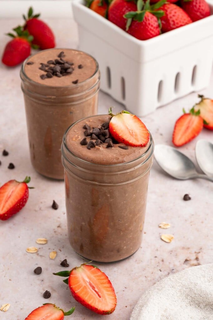 Two mason jars with chocolate overnight oats topped with mini chocolate chips and sliced strawberries.