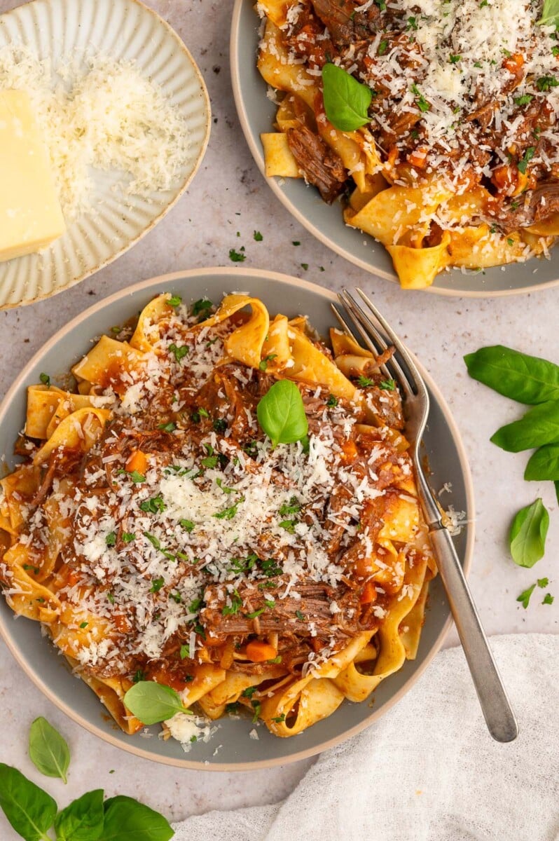 A plate of chuck roast pasta topped with cheese and fresh herbs with a fork and another plate on the side.