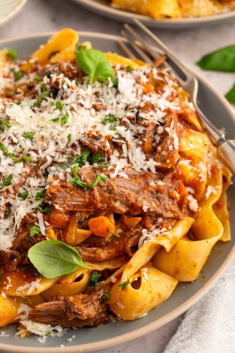 A close up of chuck roast pasta garnished with parmesan cheese and fresh herbs with a fork on the side.