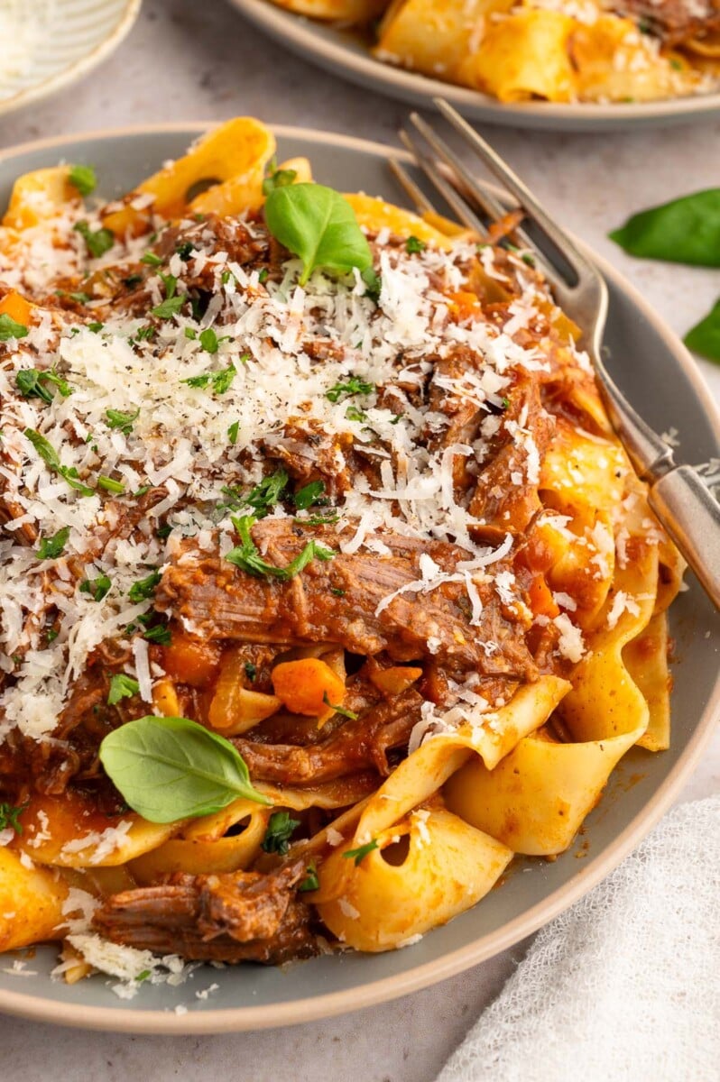A close up of chuck roast pasta garnished with parmesan cheese and fresh herbs with a fork on the side.