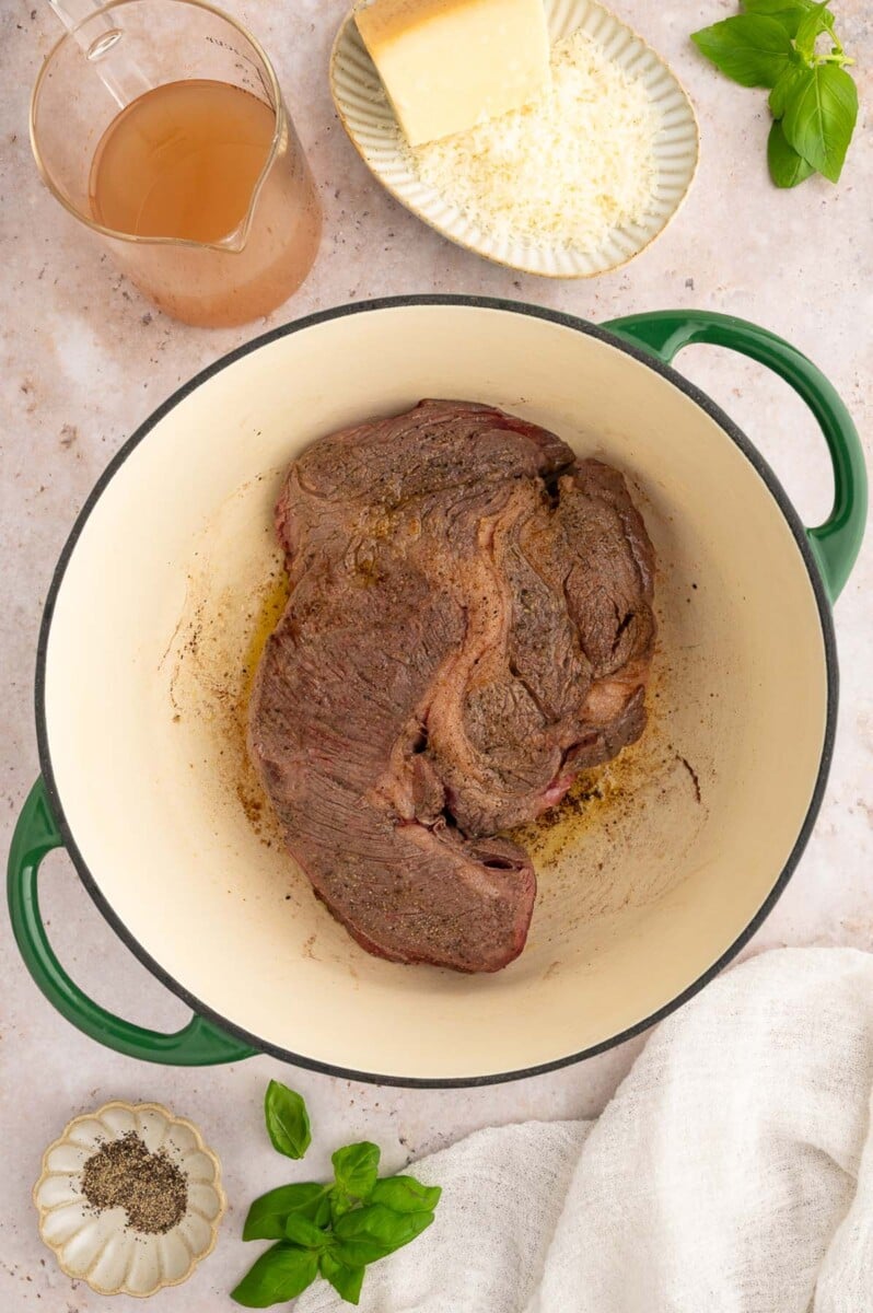 An overhead shot of a seared chuck roast in a dutch oven with beef broth and parmesan on the side.