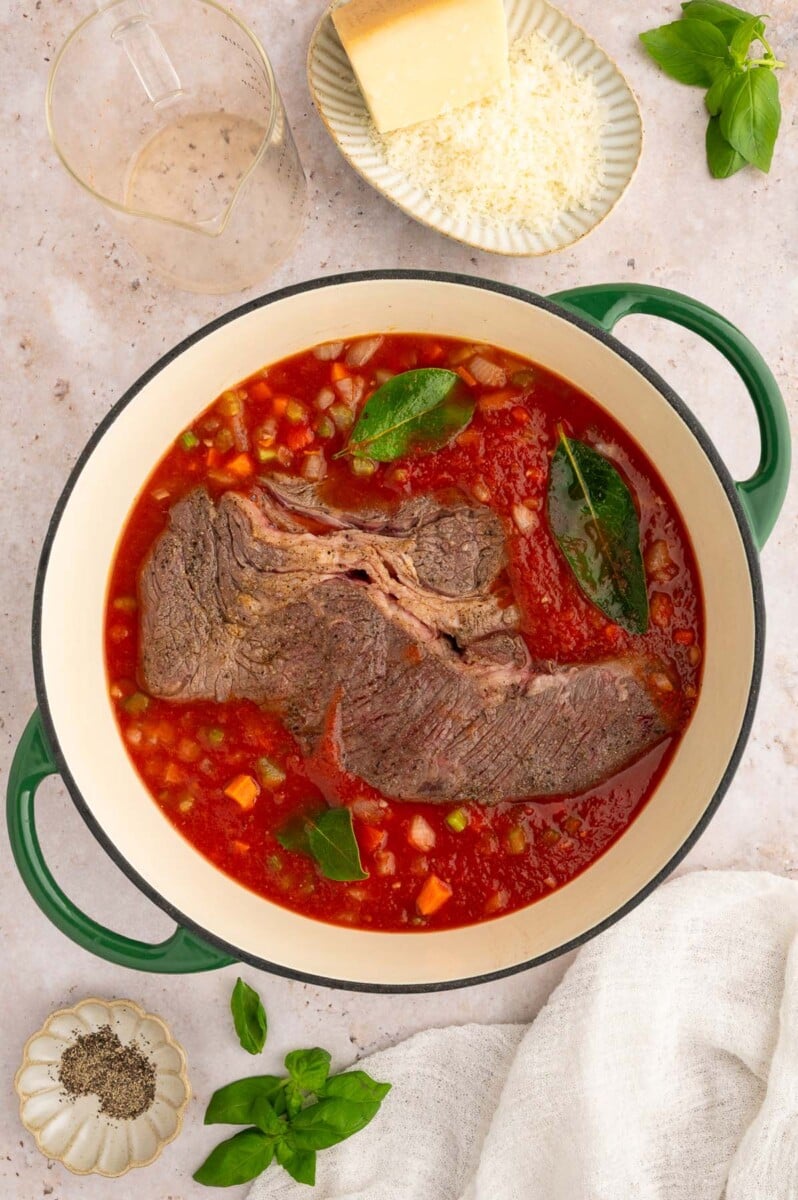A seared Chuck Roast in an uncooked pasta sauce in a dutch oven.
