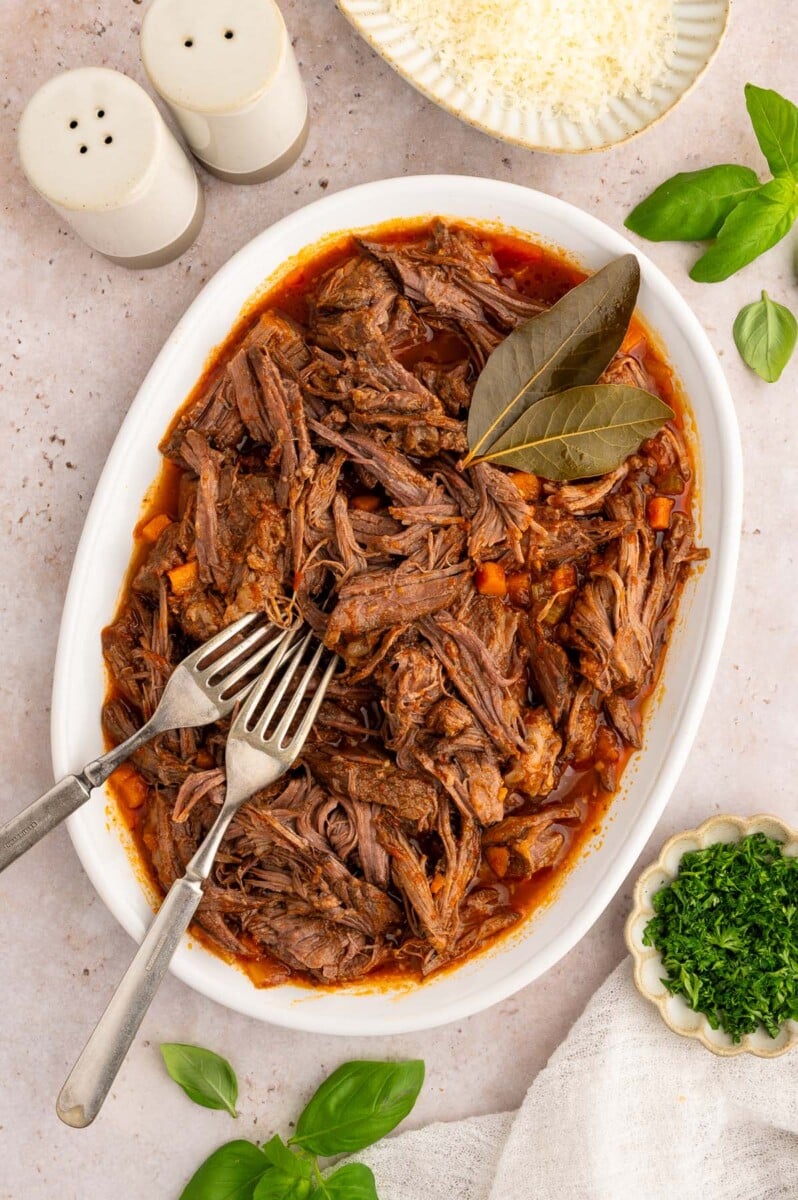 Shredded chuck roast on a white platter with two forks on it. 