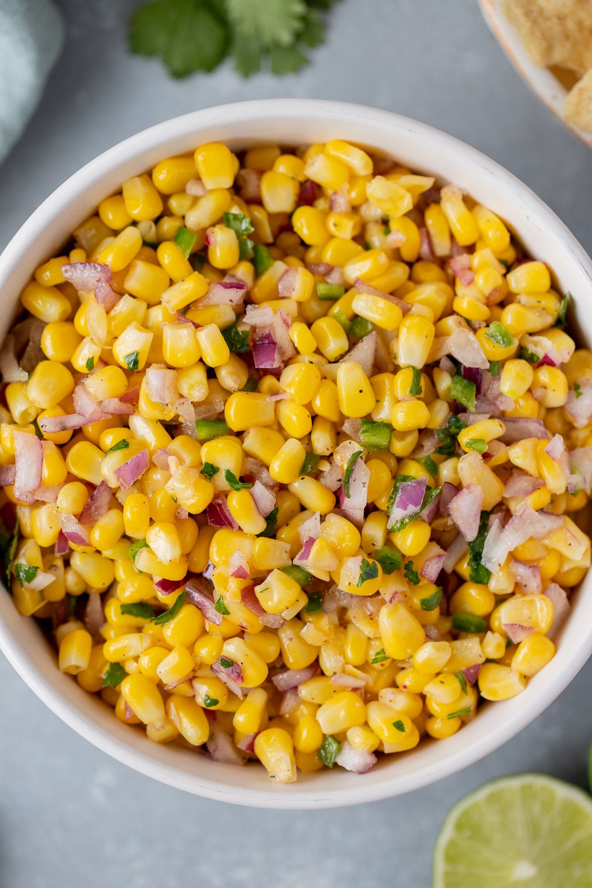 An overhead shot of a white bowl with corn salsa with a sliced lime and cilantro on the side on a grey background