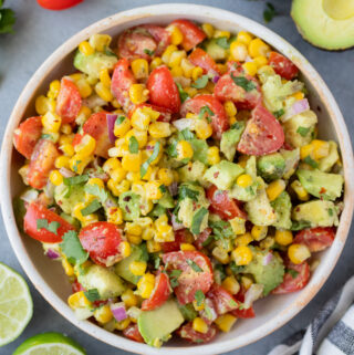 Corn Tomato Avocado Salad in a white bowl on a grey background with tomatoes, avocado and limes off to the side