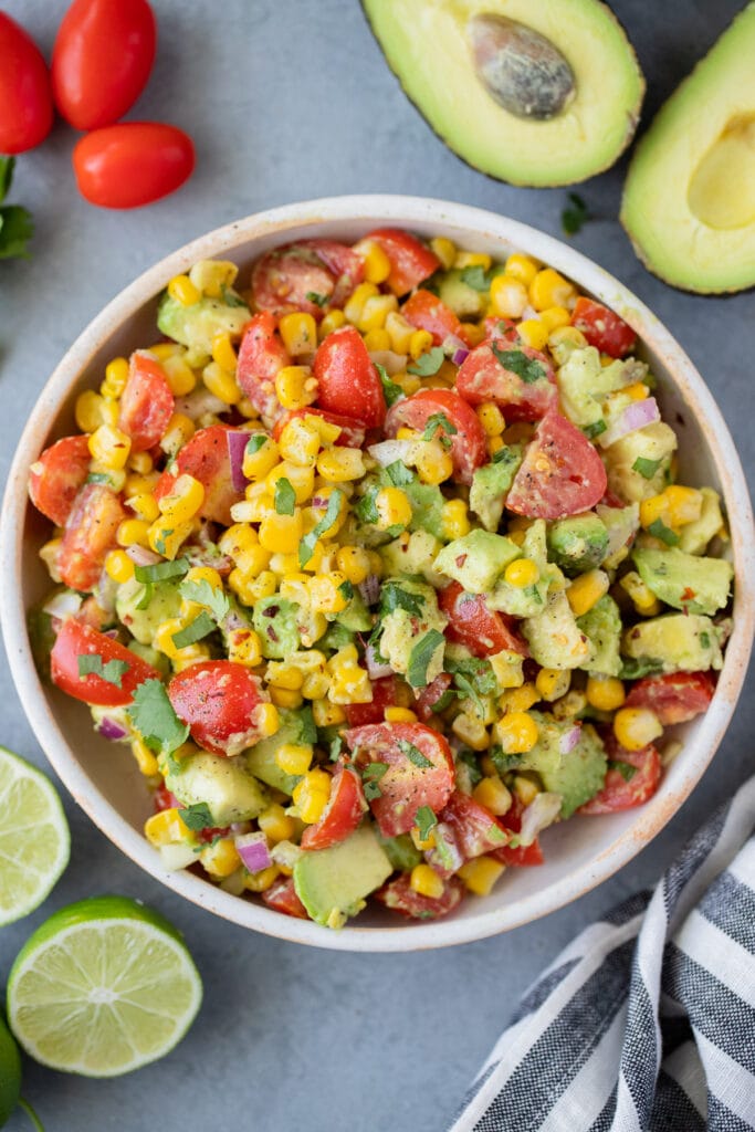 Corn Tomato Avocado Salad in a white bowl on a grey background with tomatoes, avocado and limes off to the side