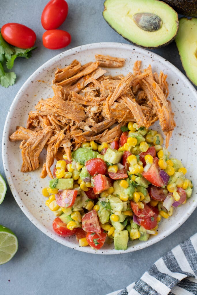 A close up of corn tomato avocado salad on a white speckled plate with pulled pork and veggies off to the side