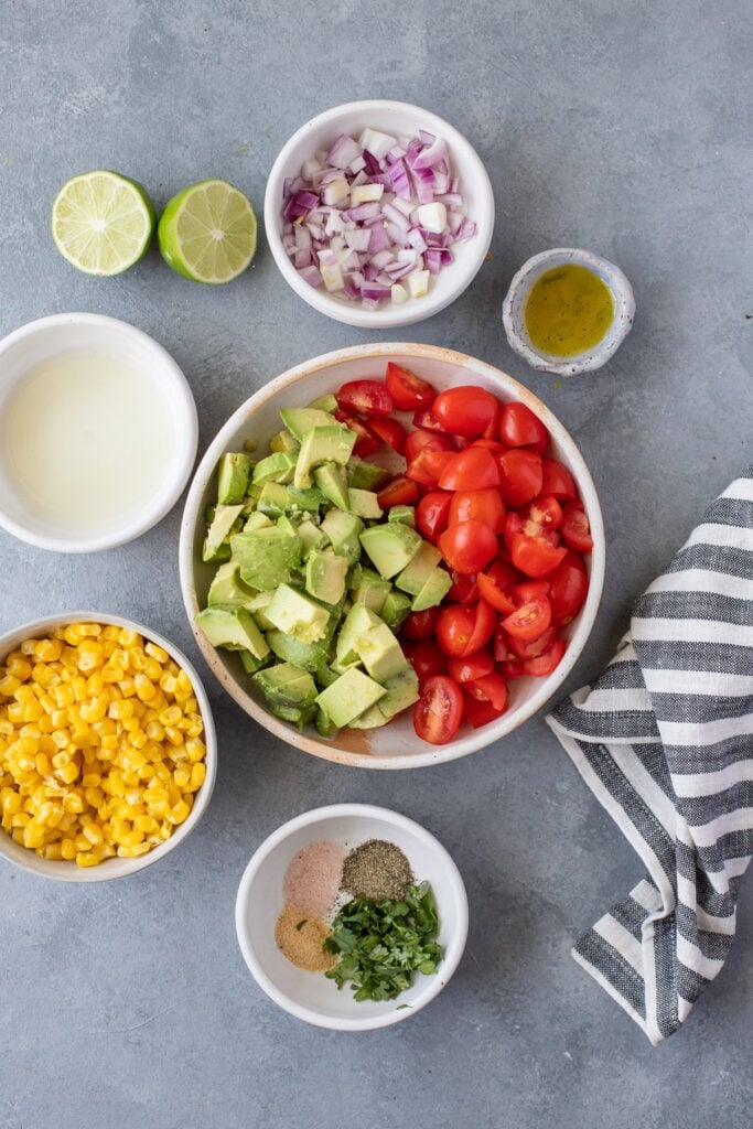 Ingredients of Corn Tomato Avocado Salad in small white bowls on a grey background: avocado, grape tomatoes, fresh corn, red onion, extra virgin olive oil, salt and pepper, garlic powder, fresh lime juice, fresh cilantro 