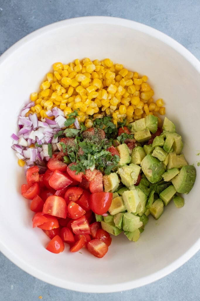 Ingredients for Corn Tomato Avocado Salad in a white bowl before being mixed together with lime juice, red onion
