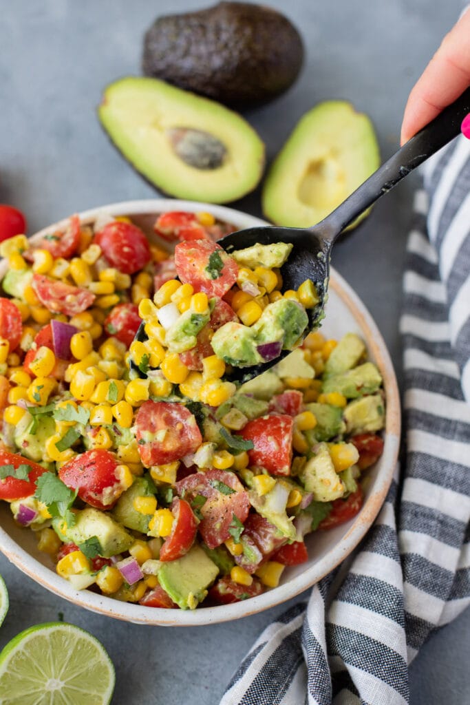A black serving spoon taking a scoop out of corn tomato avocado salad