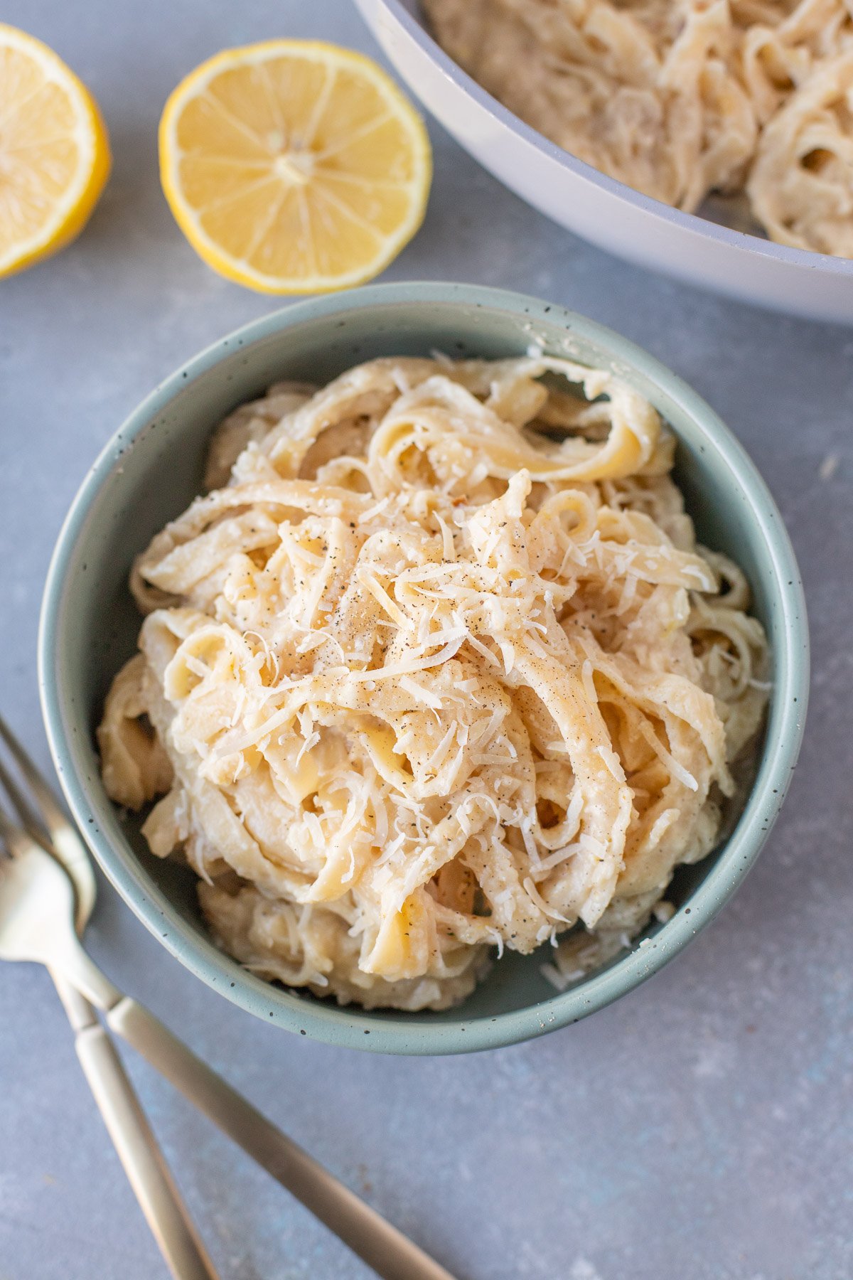 An overhead shot of a blue bowl of creamy lemon pasta with forks, a pan of pasta and lemons on the side
